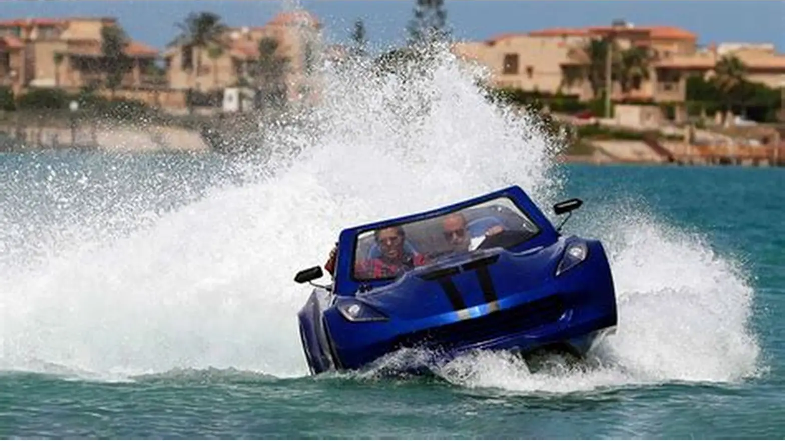 Blue car-shaped boat speeding through water, creating a large white splash with buildings behind.