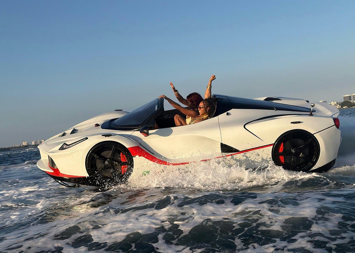 Two women wave excitedly while riding a white sports-car-shaped speedboat through splashing ocean water.