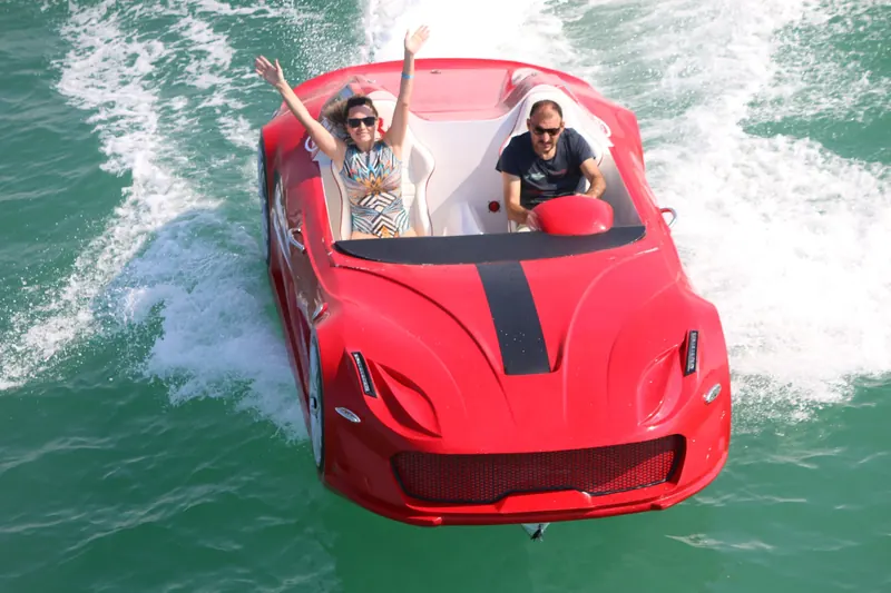 Red sports car-shaped boat speeding on turquoise water with two people enjoying the ride.